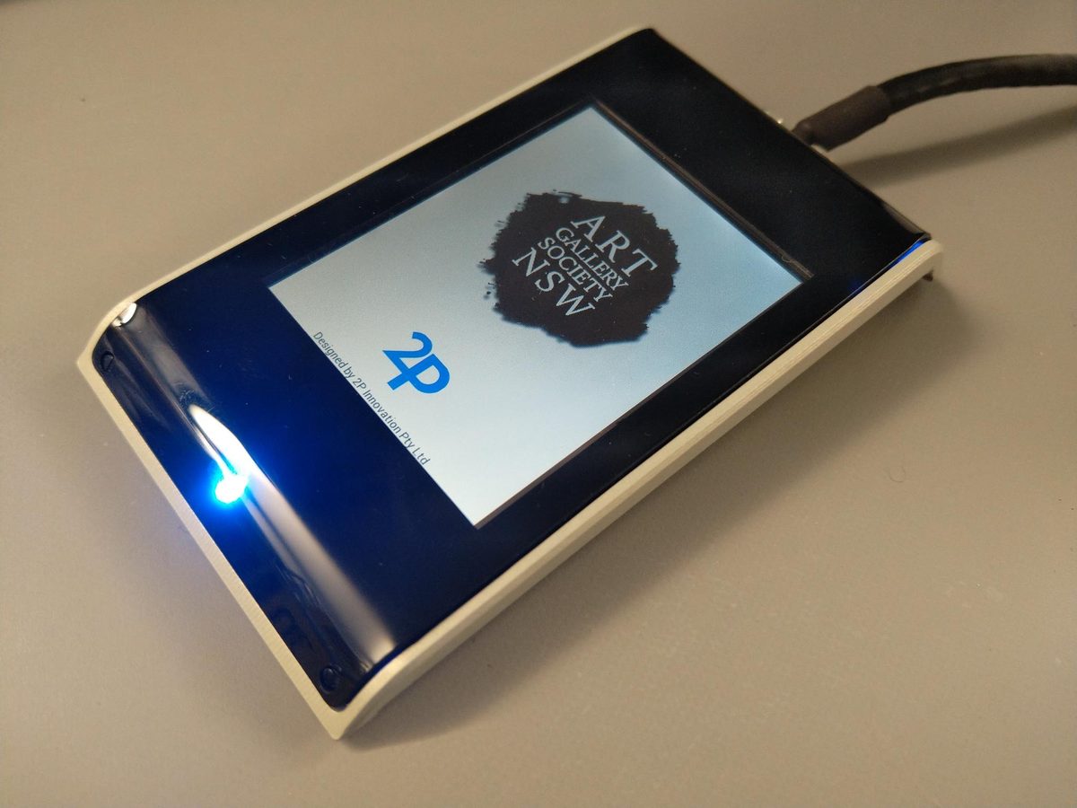 An RFID reader in a white and dark blue case, screen showing the Art Gallery Society NSW logo and the line 'Designed by 2P Innovation Pty Ltd'.