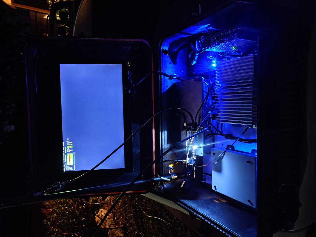 An instrument enclosure open in the field at night, interior lit blue, cables running out to equipment on the ground.