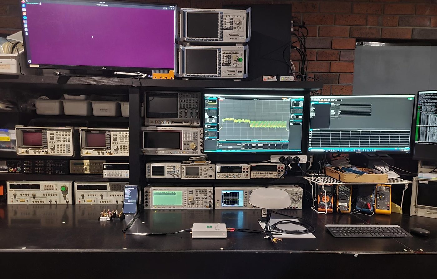 An RF test bench with a spectrum analyser, signal generators, oscilloscope, power supplies and several monitors showing measurement traces.