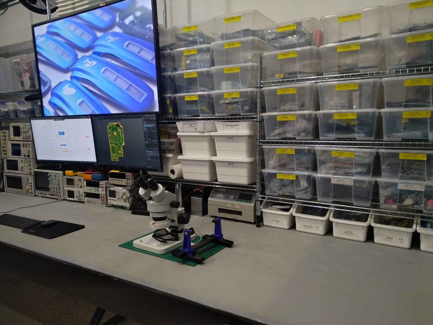 A workshop bench with a stereo inspection microscope, a monitor showing a magnified board, and racked component storage bins behind.