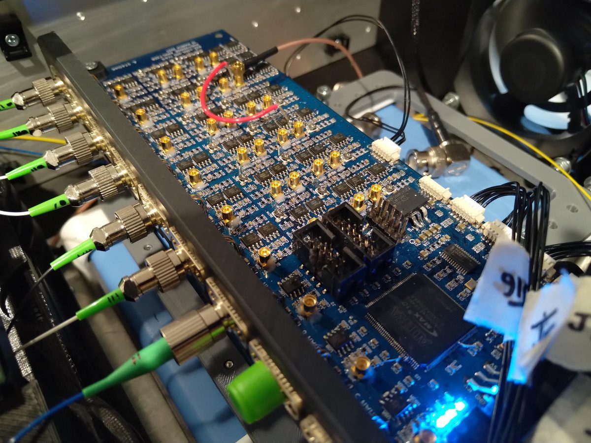 A large blue multi-channel instrument board under test, with fibre-optic and coaxial leads connected, blue indicators lit.