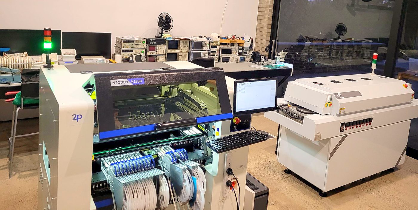 A NeoDen K1830 pick-and-place machine with feeders and component reels loaded and its status light green, beside a NeoDen T8L reflow oven, with instrument racks on the bench behind.