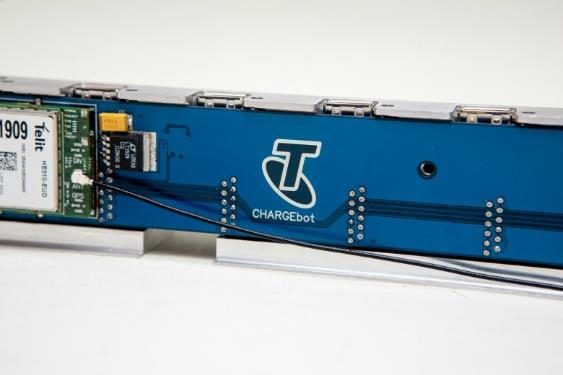 A long blue circuit board carrying Telstra CHARGEbot branding, with a Telit cellular module and rows of charging ports, mounted in an aluminium extrusion.