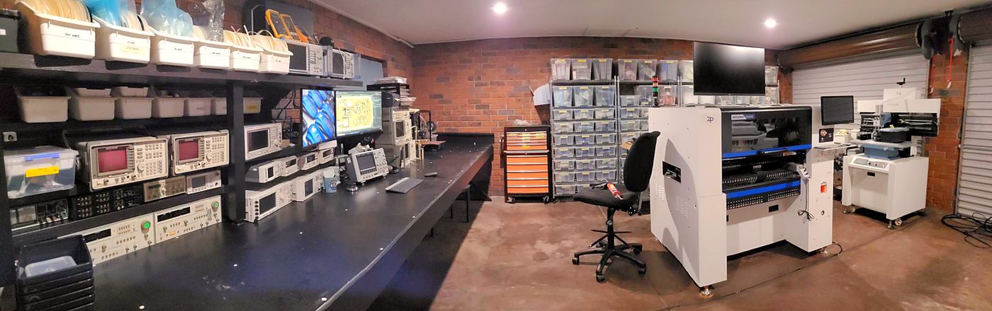 A wide view of the workshop: a long bench of spectrum analysers, signal generators and oscilloscopes along a brick wall, component storage bins, and a pick-and-place machine, stencil printer and reflow oven at the far end.